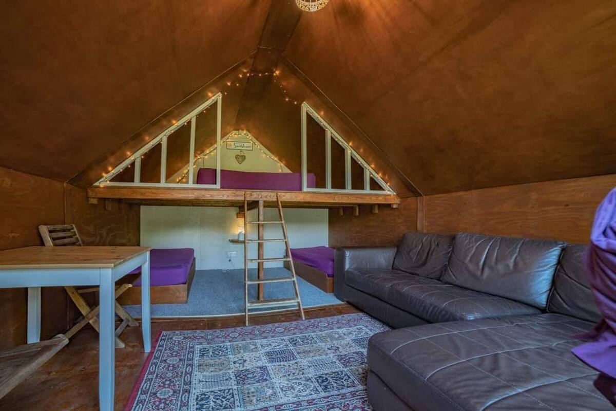 A cozy attic bedroom with a loft bed, a wooden ladder, a small table, a leather sofa, and a patterned rug.