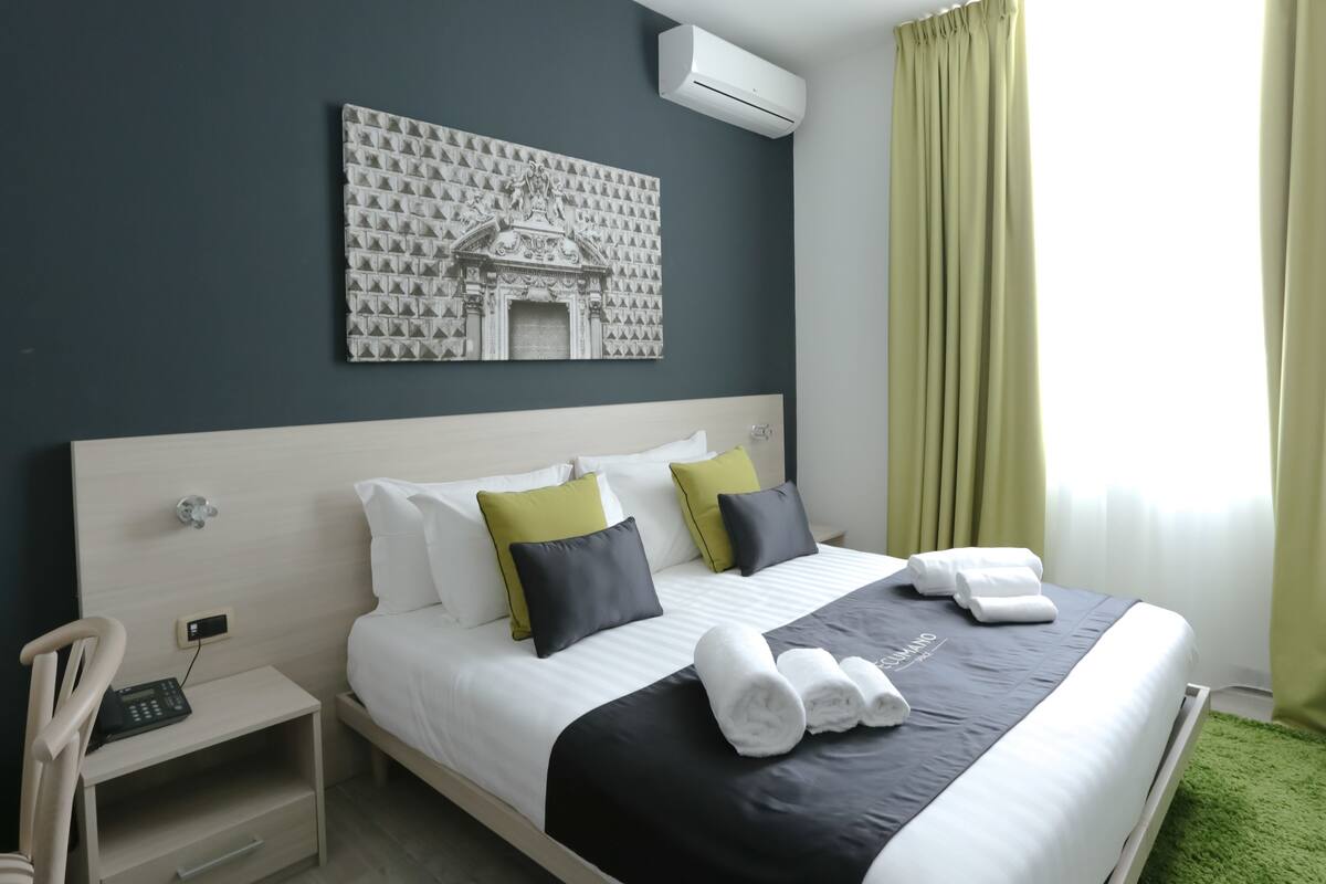 Habitación de hotel con cama, mesita de noche, una silla, aire acondicionado instalado en la pared y una ventana con cortinas.