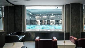 Habitación de hotel moderna con vista a una piscina interior. La habitación tiene un ventanal grande, una zona de estar con sillas y una mesita pequeña.