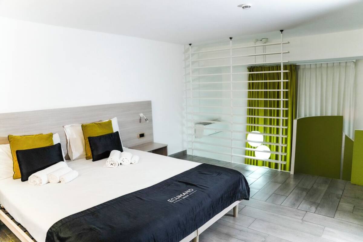 Una habitación de hotel moderna con una cama bien hecha, una mesita de noche con un teléfono y una lámpara, y un ventanal con vistas al exterior.