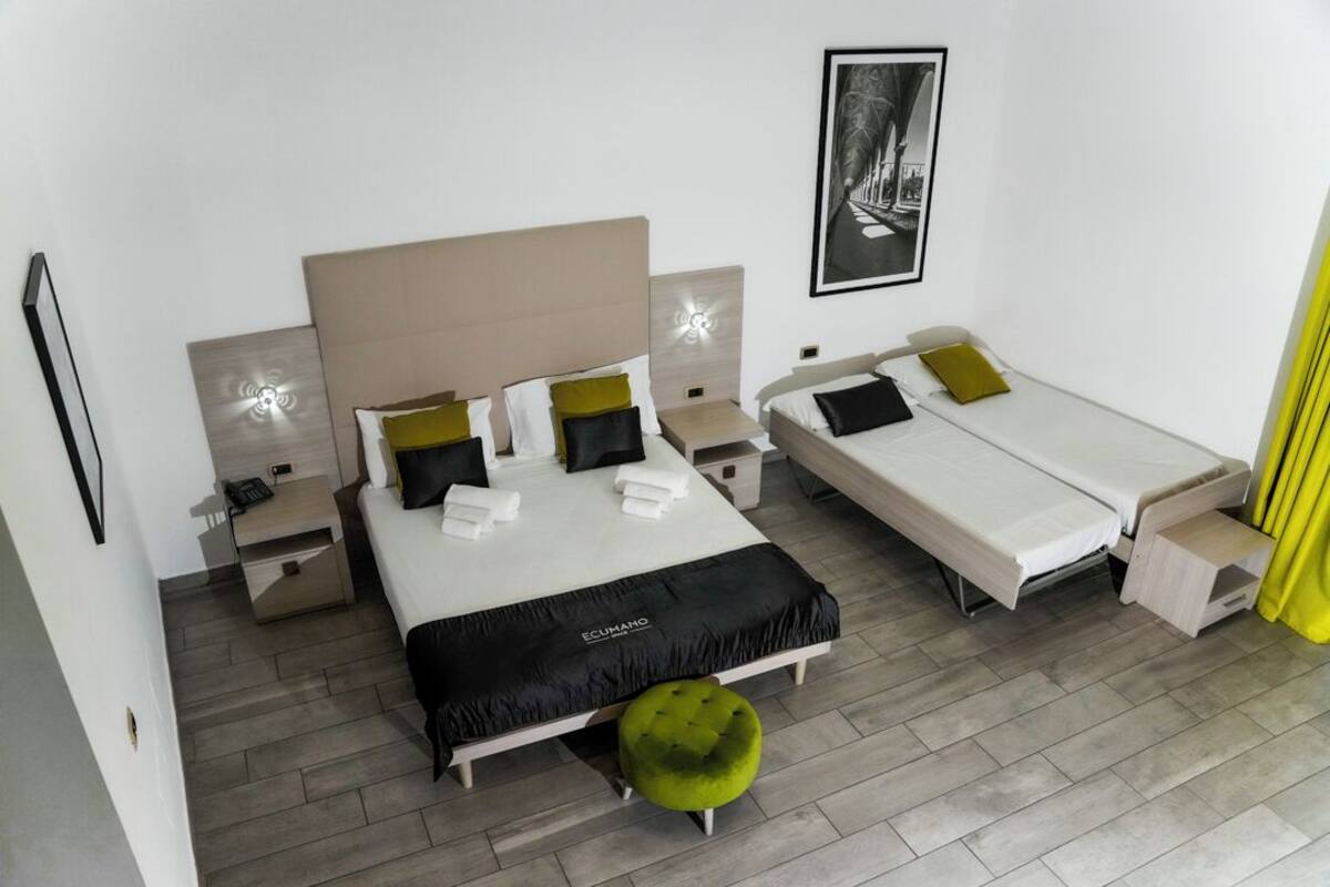Habitación de hotel con una cama grande, dos camas más pequeñas, un reposapiés verde y un cuadro enmarcado en la pared.