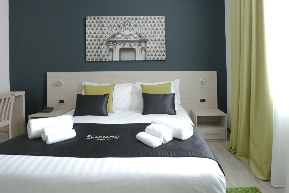Una cama bien hecha con colcha blanco y negro, dos almohadas y una toalla doblada. A la izquierda, un escritorio pequeño con una silla, y a la derecha, una mesita de noche con una lámpara.