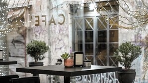 Un café al aire libre con pared de vidrio, mesas, sillas y luces decorativas.