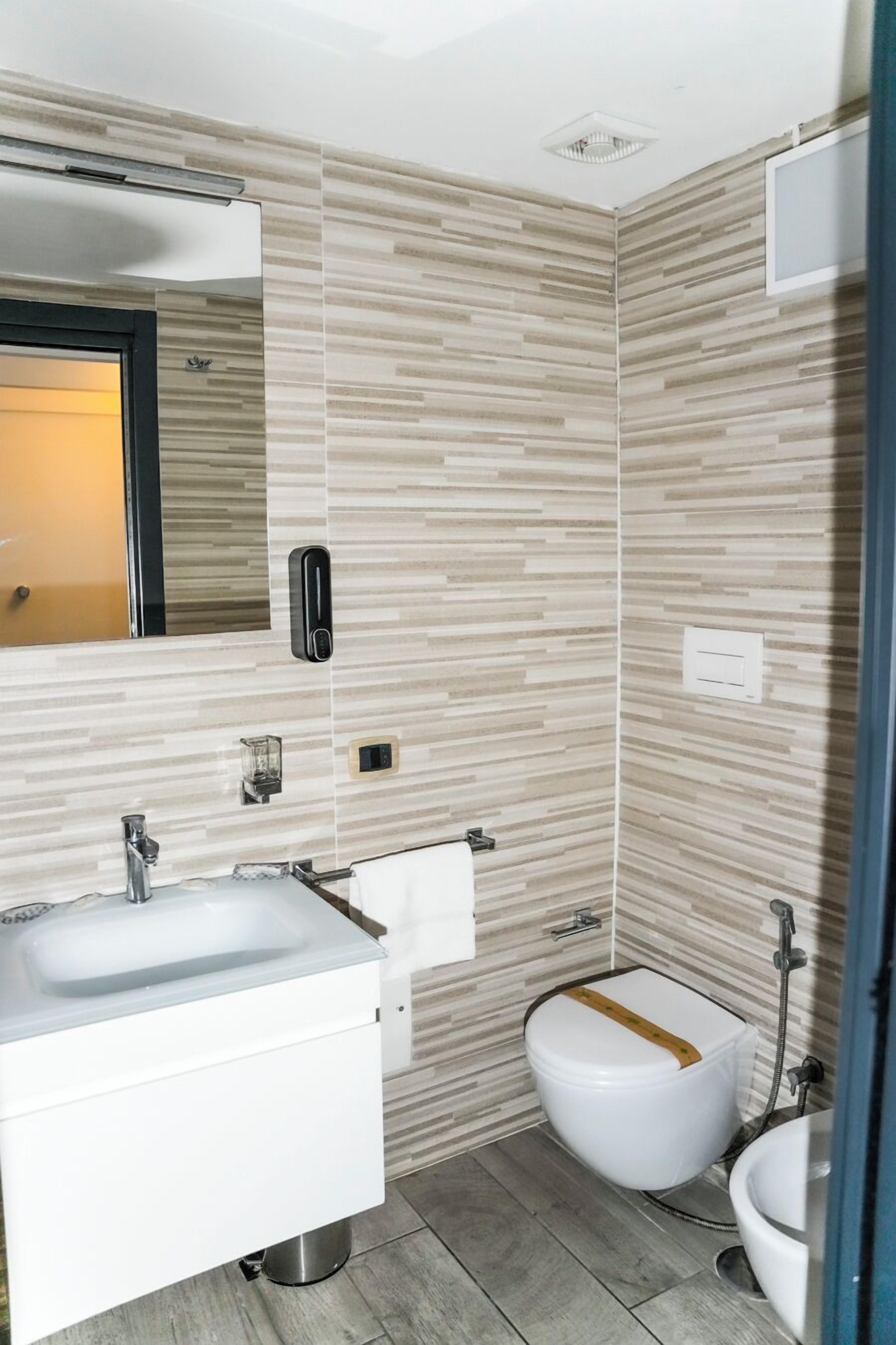 Un baño moderno con lavamanos, espejo y váter blancos. Las paredes tienen un diseño que imita la madera, y a la derecha se encuentra la zona de ducha.