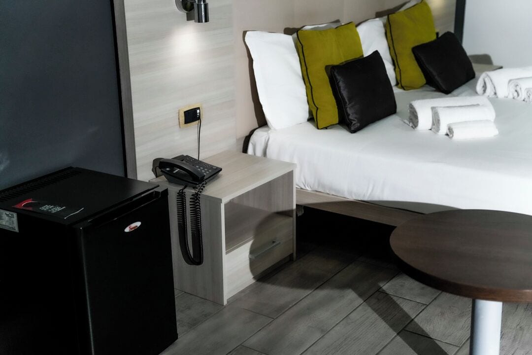 Camera d'albergo con un letto, una scrivania con il telefono, un piccolo frigorifero nero e un tavolino rotondo in legno.