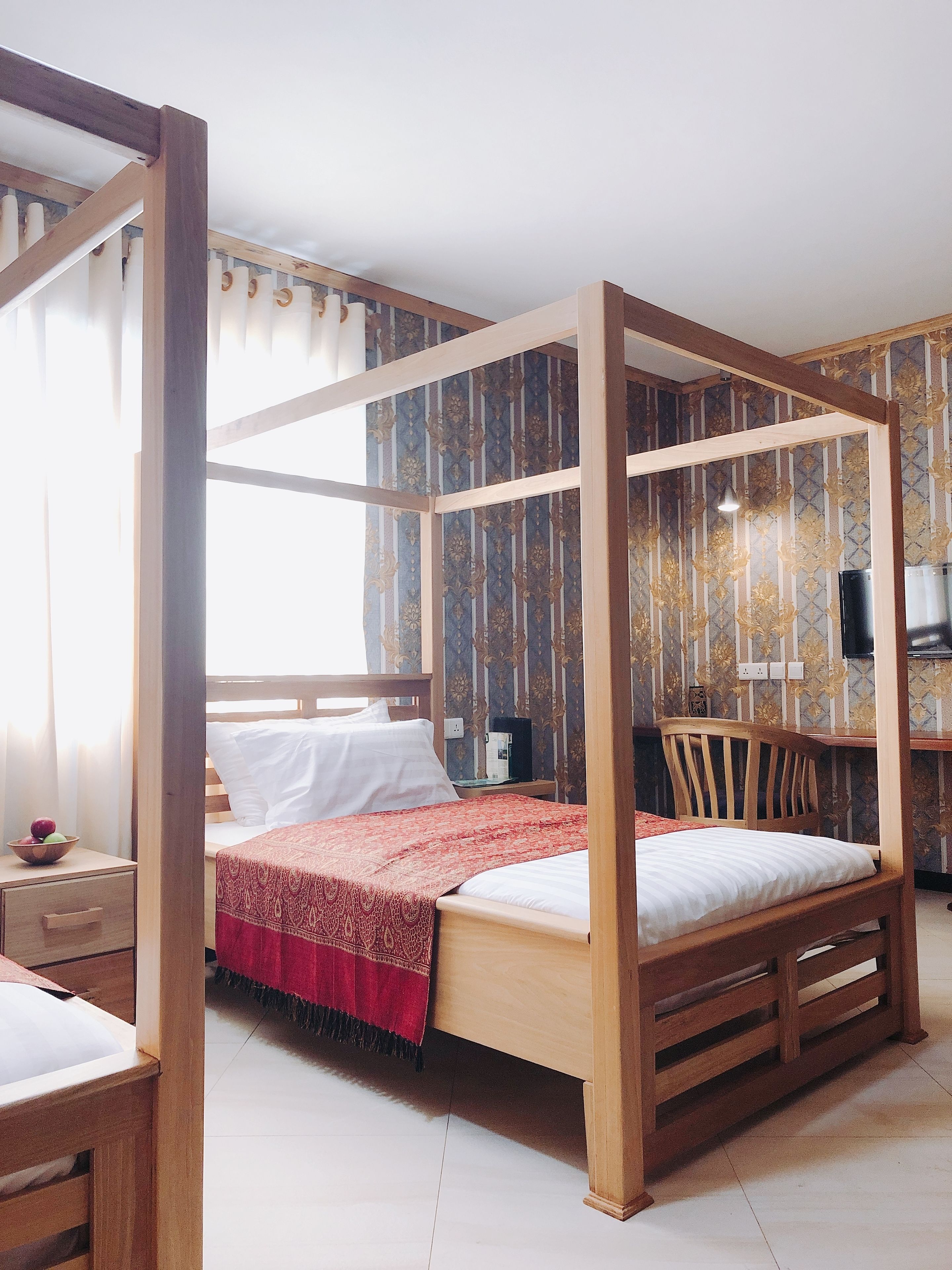 Una cama con dosel de madera y colcha roja y blanca. Hay una mesita de noche de madera con un frutero y una lámpara. La habitación tiene papel tapiz estampado y una silla de madera.