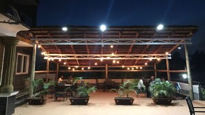 Un patio al aire libre con mesas y sillas de madera, plantas en macetas y una zona de estar cubierta con luces colgantes.