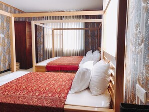 Una habitación con dos camas, una con colcha roja y la otra con ropa de cama blanca. Una de las camas tiene cabecero de madera, y la habitación presenta papel tapiz estampado.