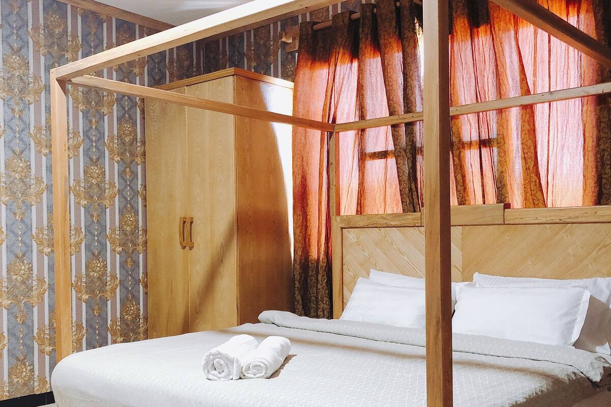 A four-poster bed with a wooden frame, white linens, and a red patterned rug.