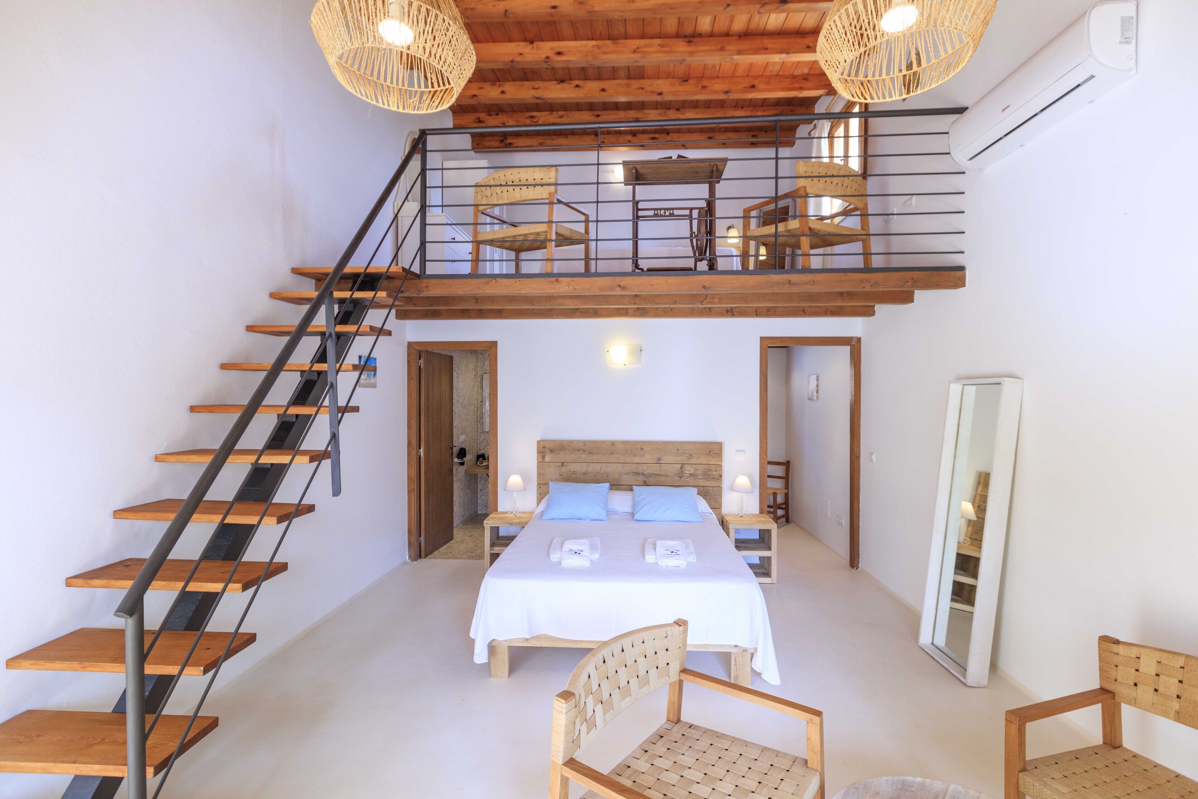 A bedroom with a large bed, a wooden staircase, and a balcony with wicker furniture.