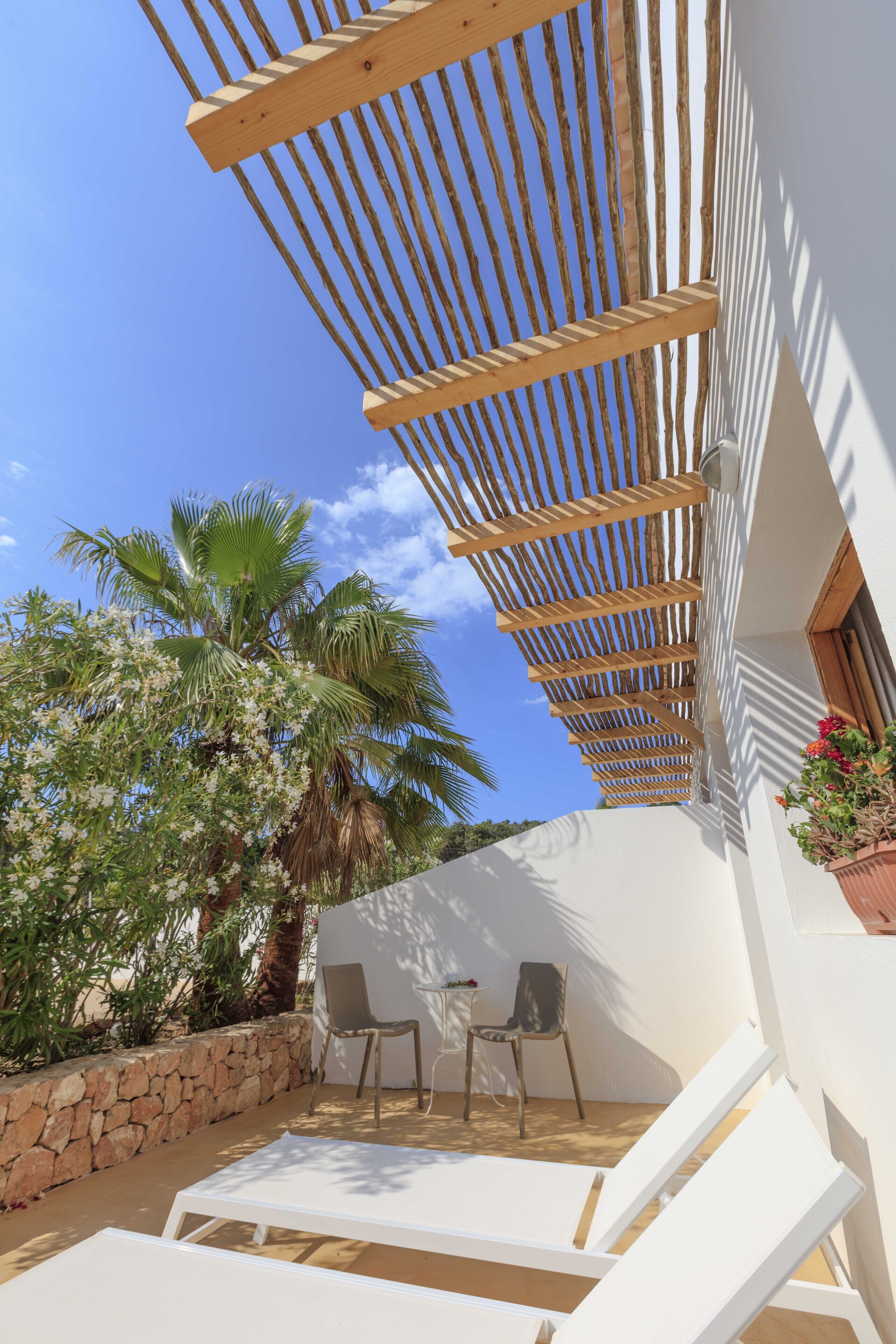 A covered outdoor seating area with white lounge chairs, a stone wall, and a wooden pergola.