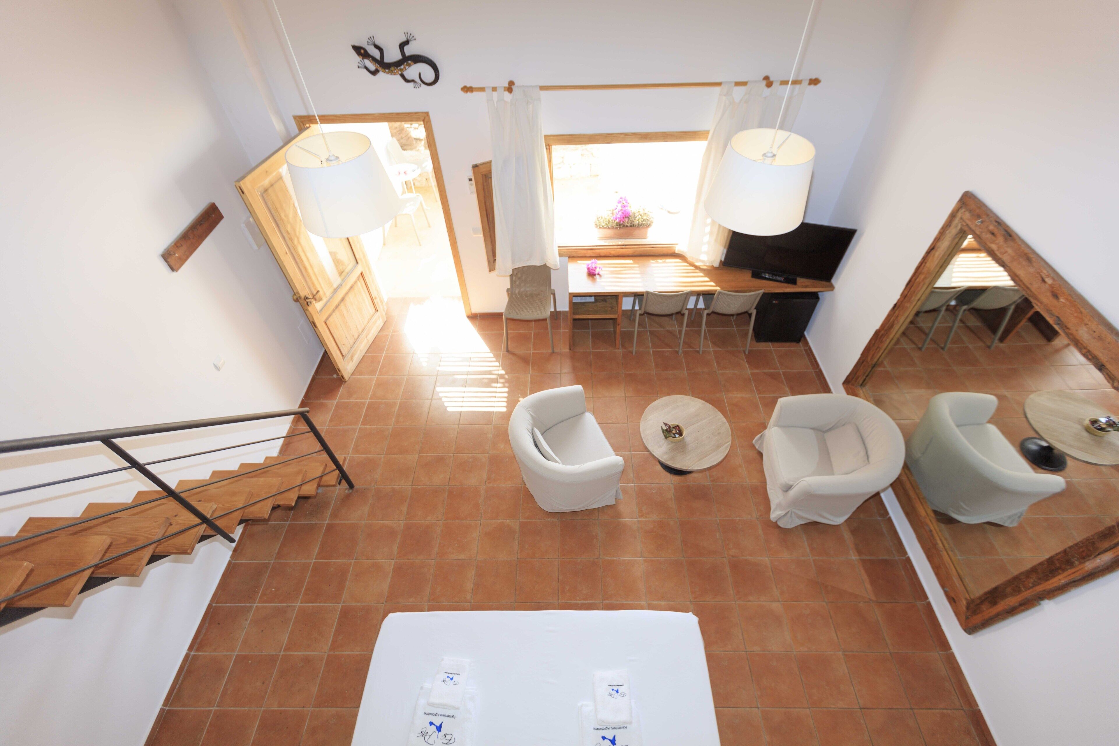 Una habitación amplia con comedor, dos sillones blancos, una mesita redonda, un espejo grande y una escalera.