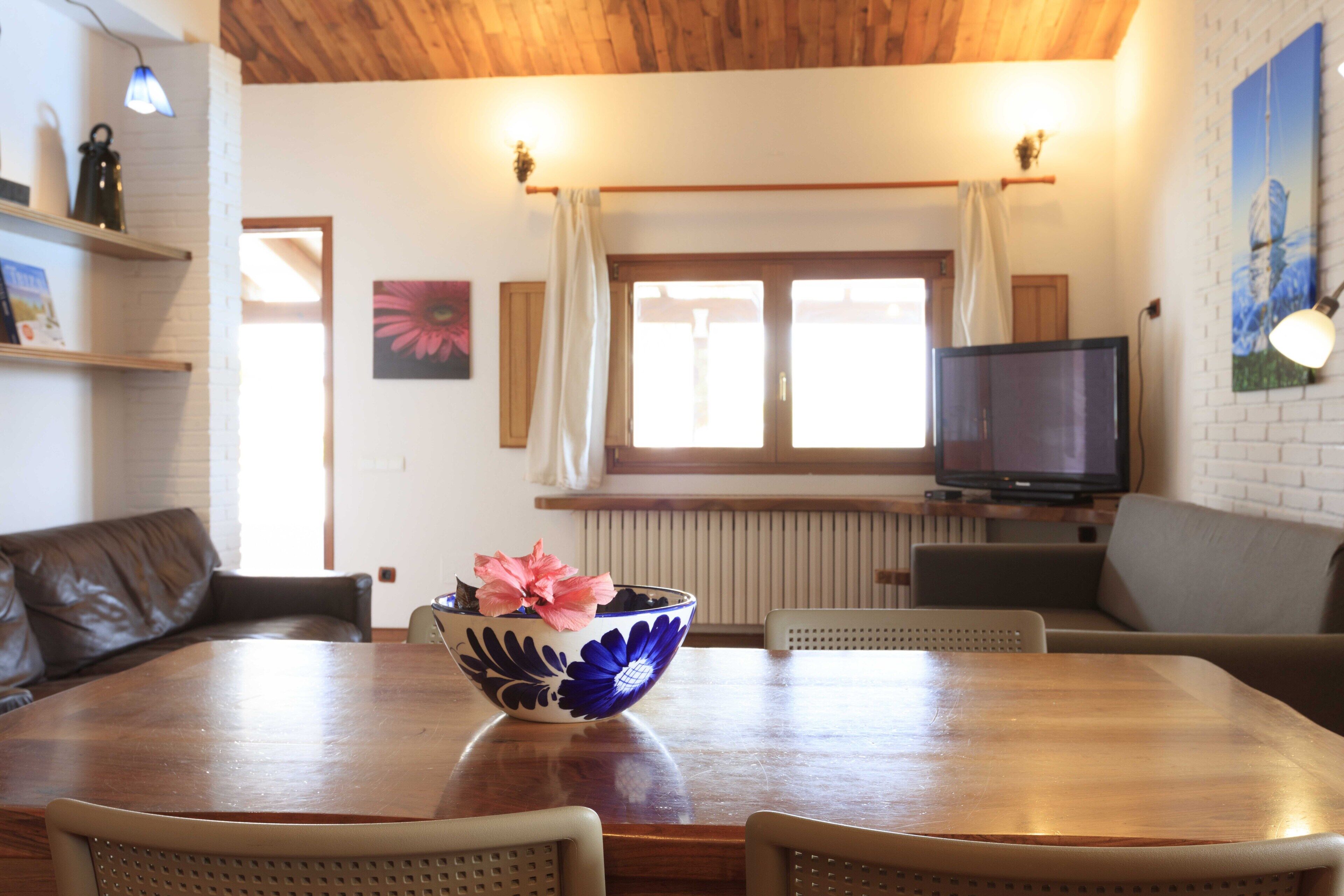 A dining area with a wooden table, chairs, a flower bowl, a television, and a window with curtains.