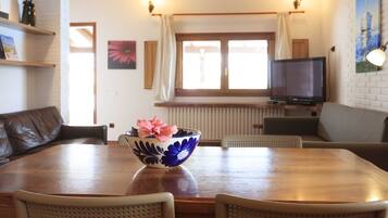 A dining area with a wooden table, chairs, a flower bowl, a television, and a window with curtains.