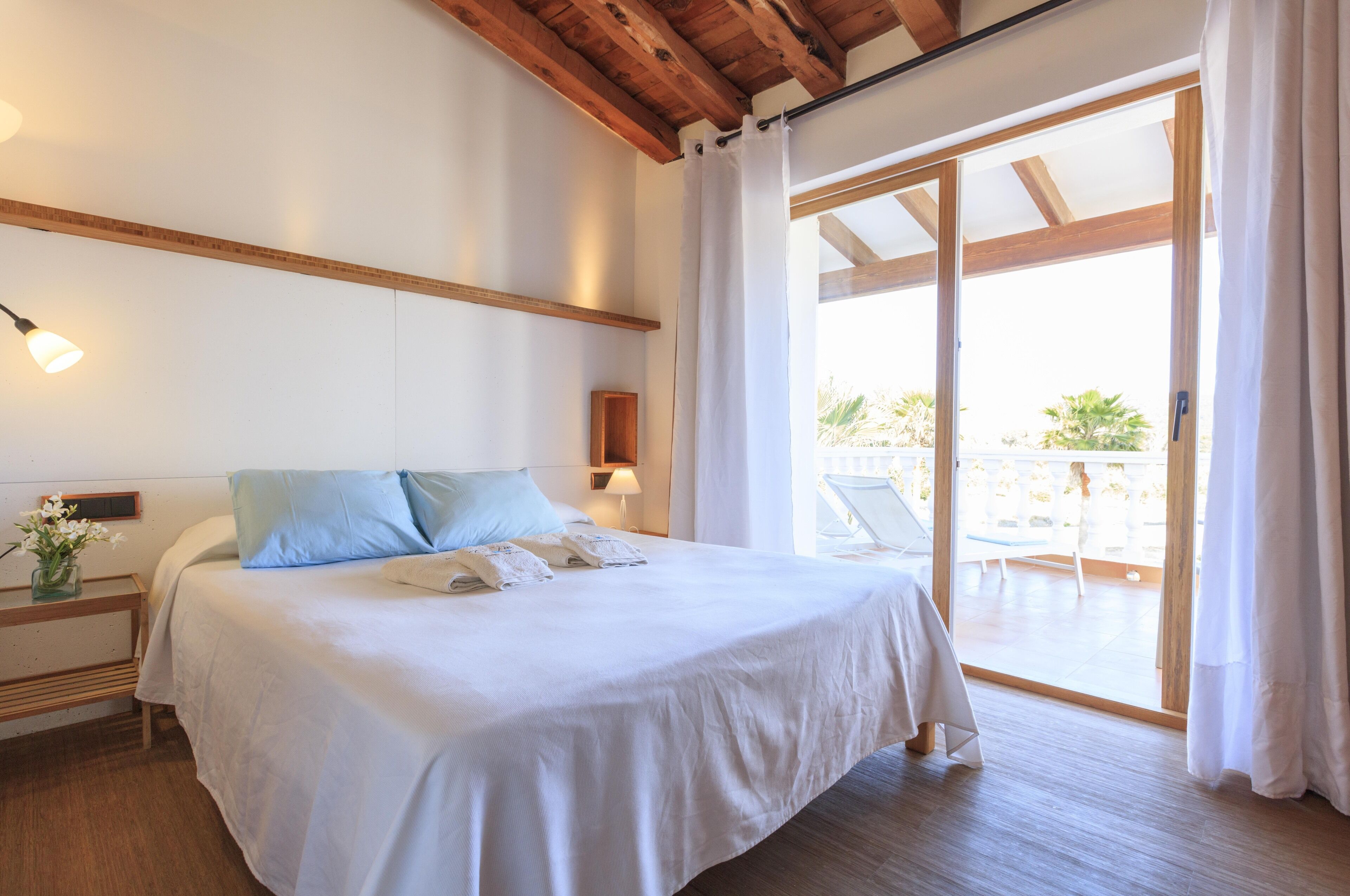 A neatly made bed with pillows, a bedside table with a lamp, and a view of an outdoor balcony.