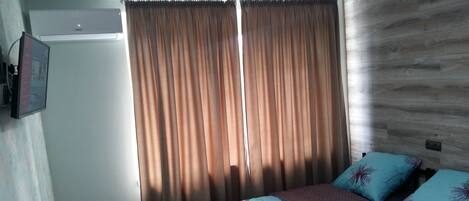 A bedroom with a bed, two pillows, a window with curtains, and a wall-mounted air conditioner.