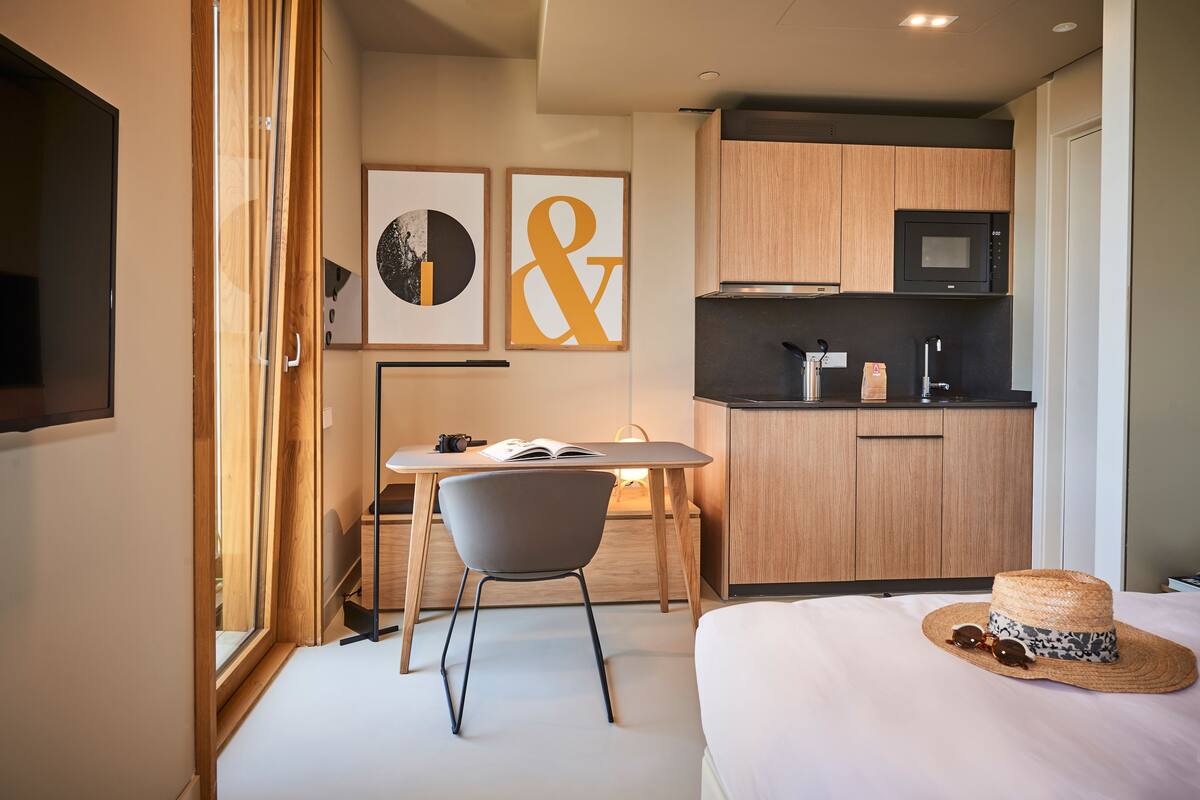 A modern hotel room with a bed, a desk with a chair, a small kitchenette, and artwork on the wall.