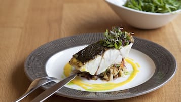 Un plato de comida con un filete de pescado, adornado con vegetales verdes, sobre un plato blanco con borde gris.