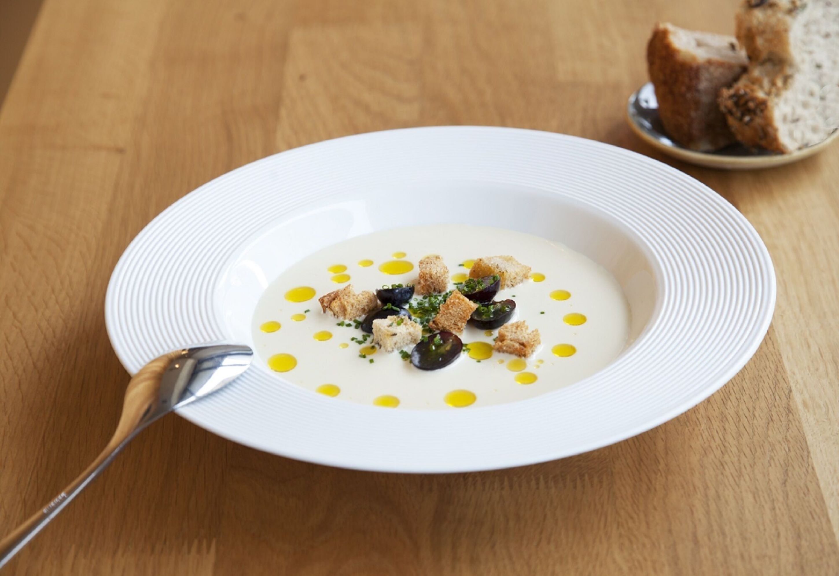 Un plato blanco con sopa cremosa adornado con croutons y aceitunas, acompañado de una cuchara, sobre una mesa de madera.