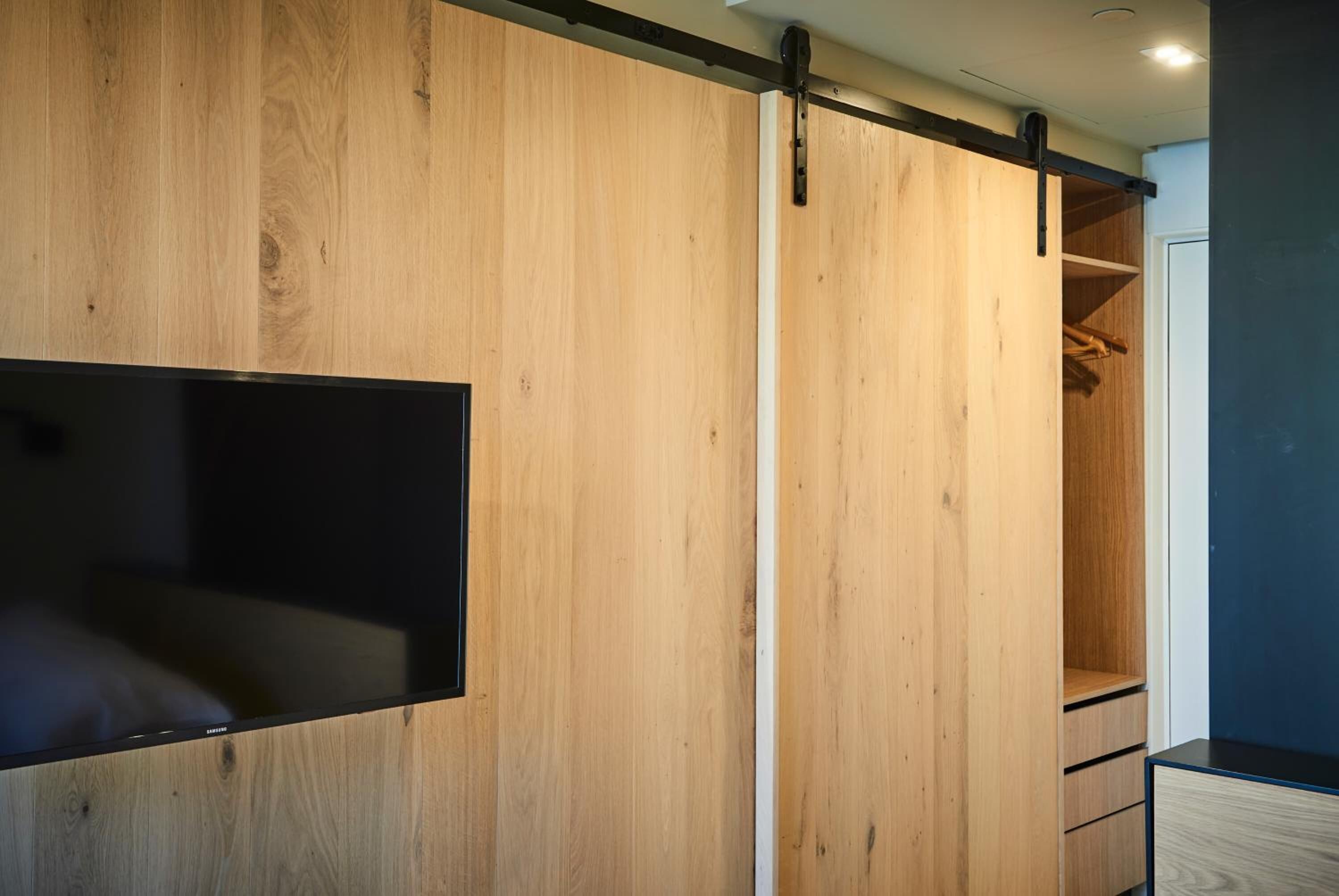 Ein Zimmer mit einem schiebbaren Holzschrank, einem schwarzen Flachbildfernseher an der Wand und einer kleinen Holzkommode.