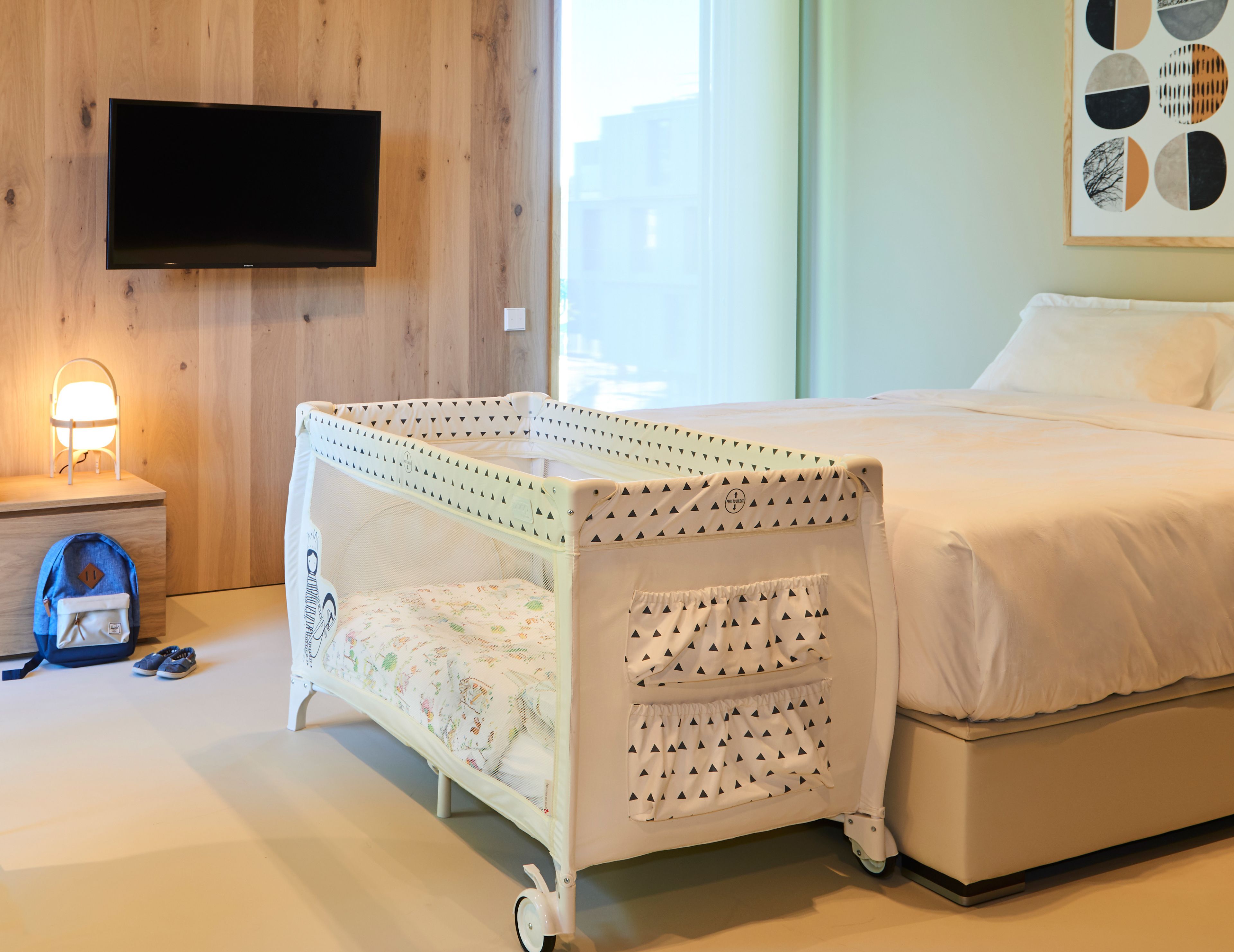 A bedroom with a crib, a bed, a TV mounted on the wall, and a bedside table with a lamp.