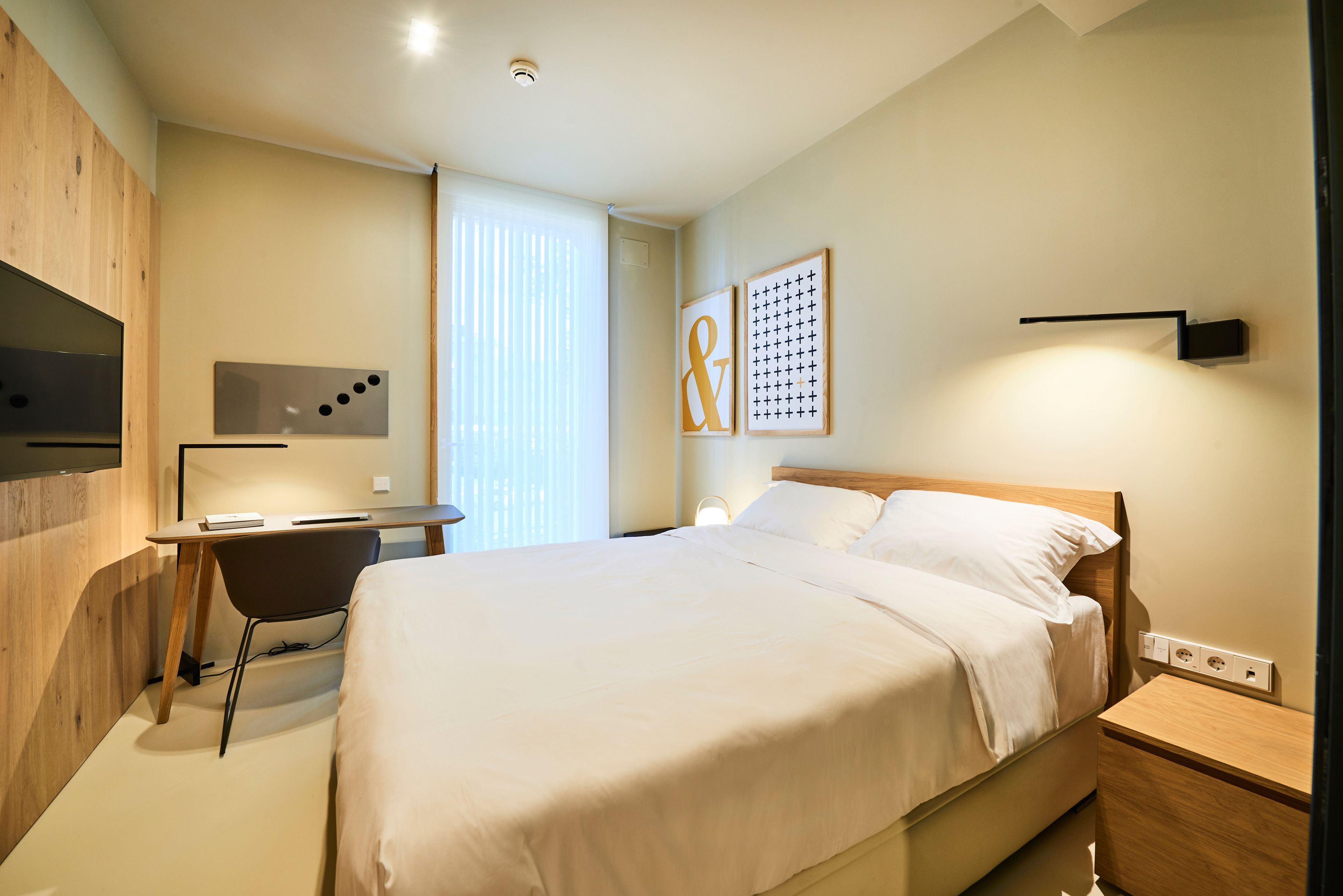 Una moderna camera d'hotel con un letto, una scrivania e una sedia. Si vede una finestra con persiane, una TV a muro e un'opera d'arte appesa al muro.