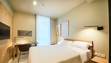 Una moderna camera d'hotel con un letto, una scrivania e una sedia. Si vede una finestra con persiane, una TV a muro e un'opera d'arte appesa al muro.