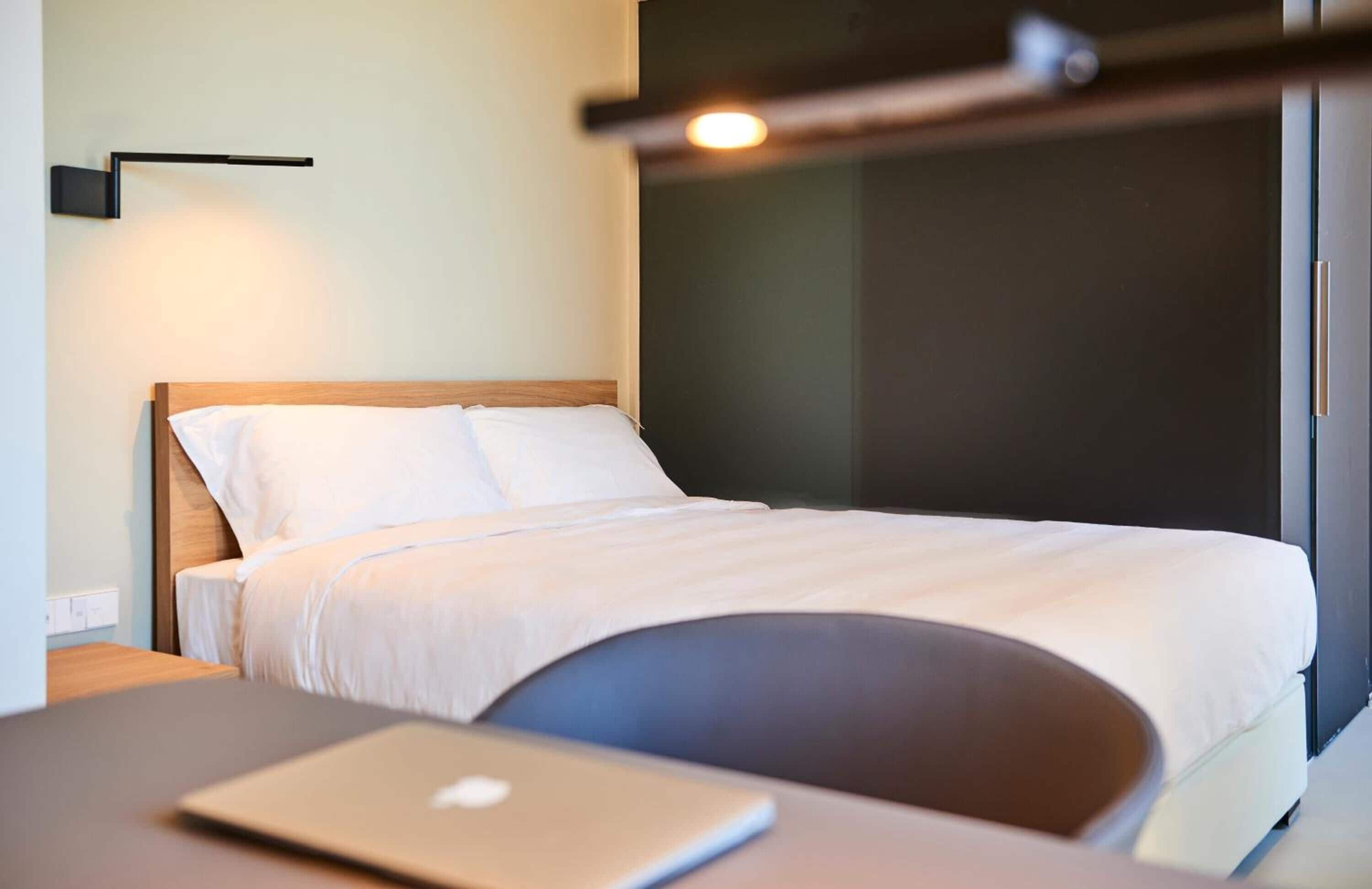 A modern hotel room with a neatly made bed, a laptop on a desk, and a wall-mounted light fixture.