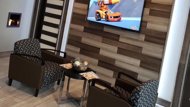 Een modern interieur met een houten wandpaneel, een flatscreen televisie waarop een auto te zien is, en een zithoek met twee gestreepe stoelen en een kleine tafel.