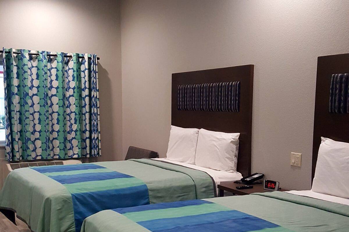 Une chambre d’hôtel avec deux lits, chacun recouvert d’une couvre-lit à rayures vertes et bleues. On y trouve une fenêtre avec un rideau, une table de chevet avec un téléphone et un réveil, et une tête de lit fixée au mur.