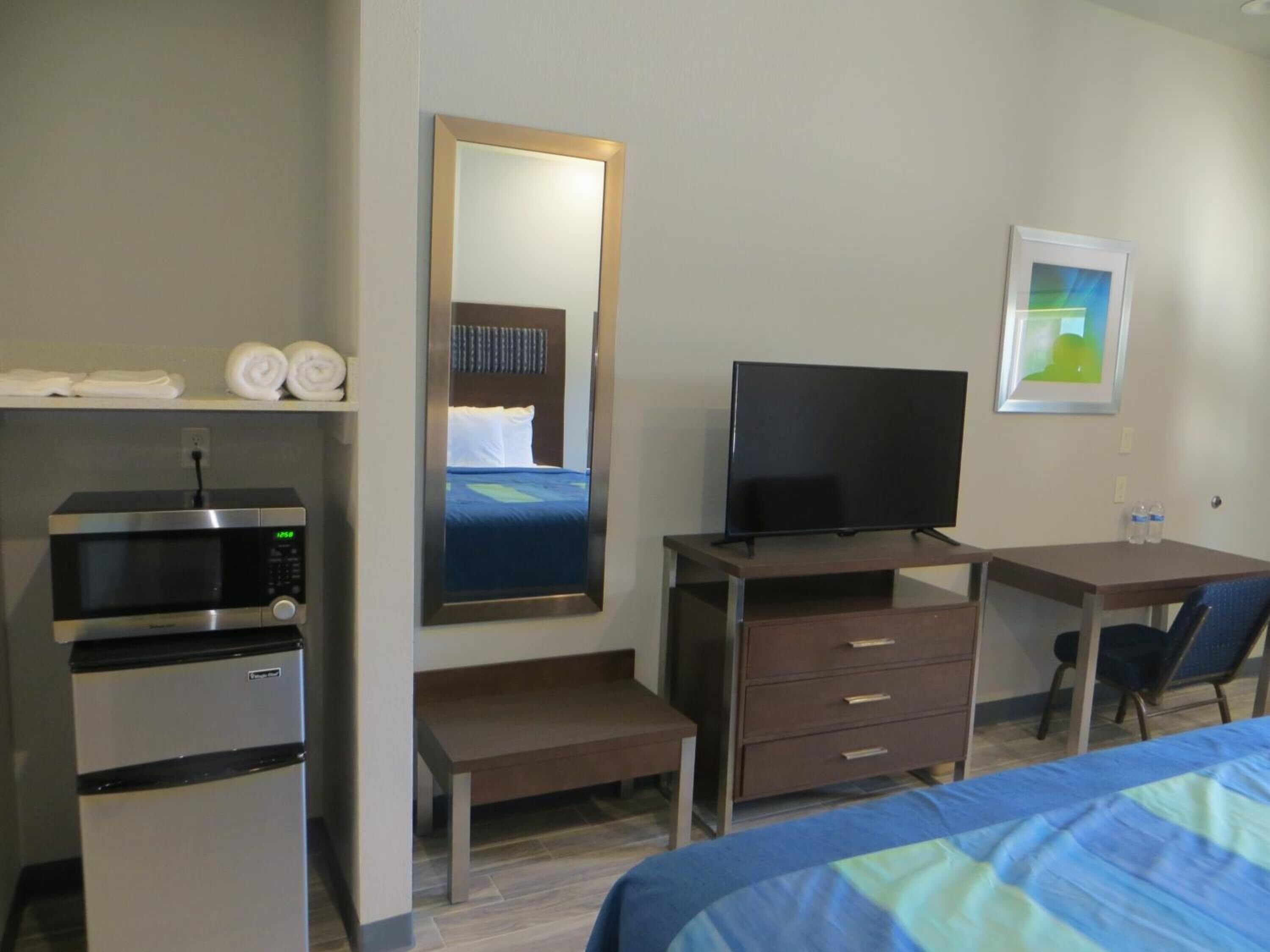 Quarto de hotel com cama, escrivaninha com televisão, micro-ondas, mini-geladeira, espelho e um quadro na parede.