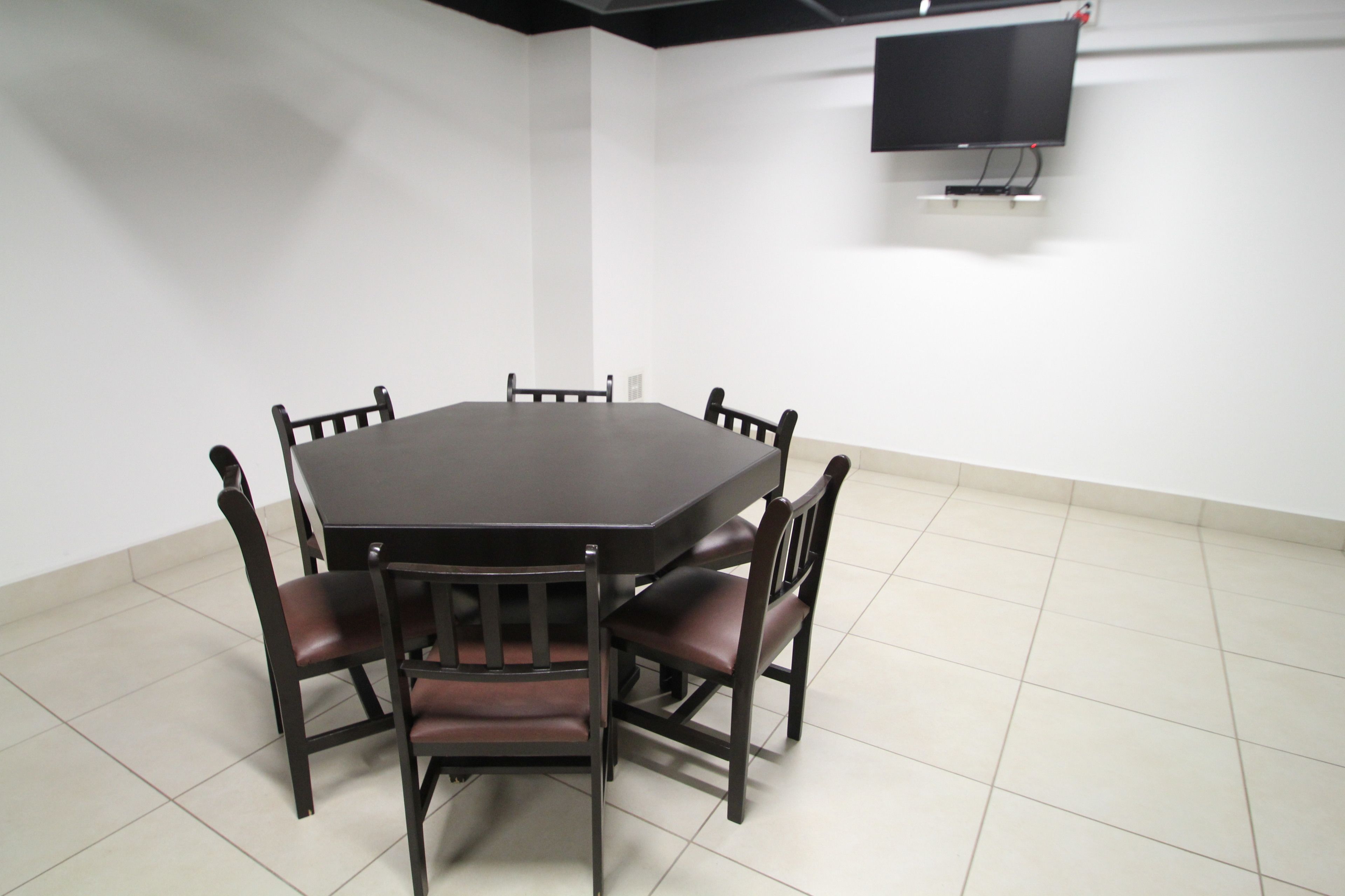 A conference room with a large table and six chairs, a flat-screen TV mounted on the wall, and a tiled floor.