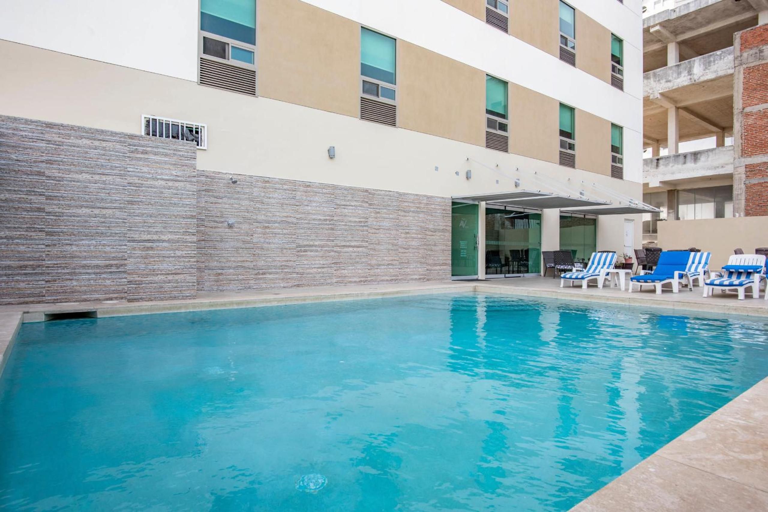 Una piscina con sillones de descanso y un área techada, rodeada por un edificio de varios pisos y ventanas.