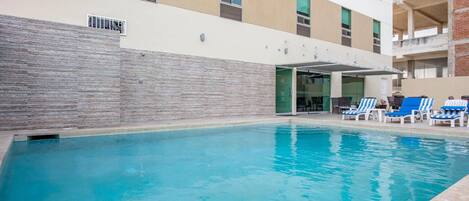 Una piscina con sillones de descanso y un área techada, rodeada por un edificio de varios pisos y ventanas.