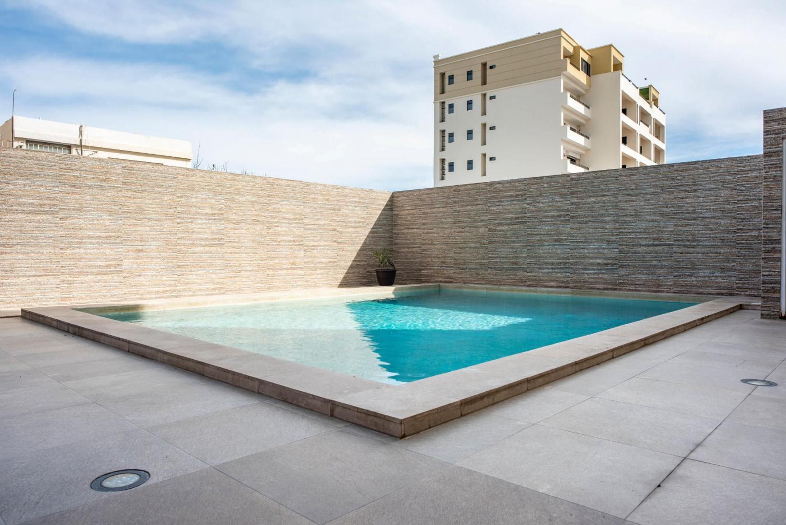 A rooftop swimming pool with a clear blue water surface, surrounded by a low brick wall and a modern building in the background.