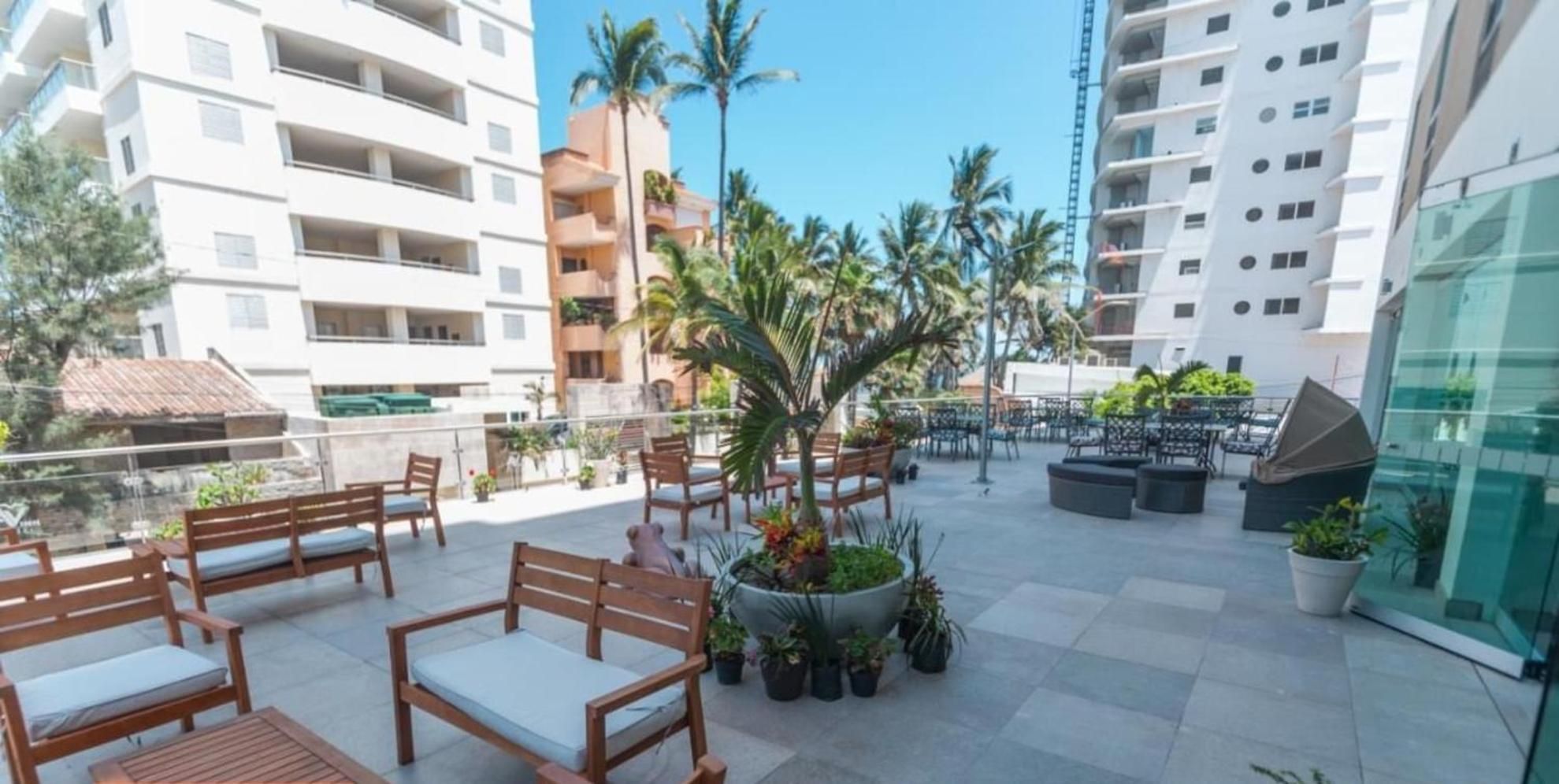 Una terraza en la azotea con mobiliario exterior, plantas en macetas y vistas de edificios circundantes.