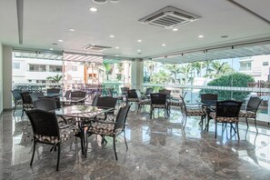 A spacious indoor dining area with multiple tables and chairs, large windows offering views of palm trees and a pool, and a ceiling with recessed lighting. - Hotel Ave Inn Mazatlán (Mazatlán)