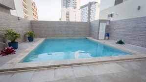 A rectangular swimming pool with a tiled deck, surrounded by a low stone wall and potted plants. - Hotel Ave Inn Mazatlán (Mazatlán)
