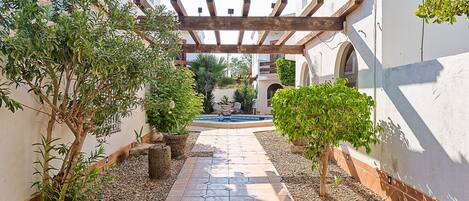 A paved pathway leading to a building with a wooden pergola, surrounded by potted plants and a small fountain.