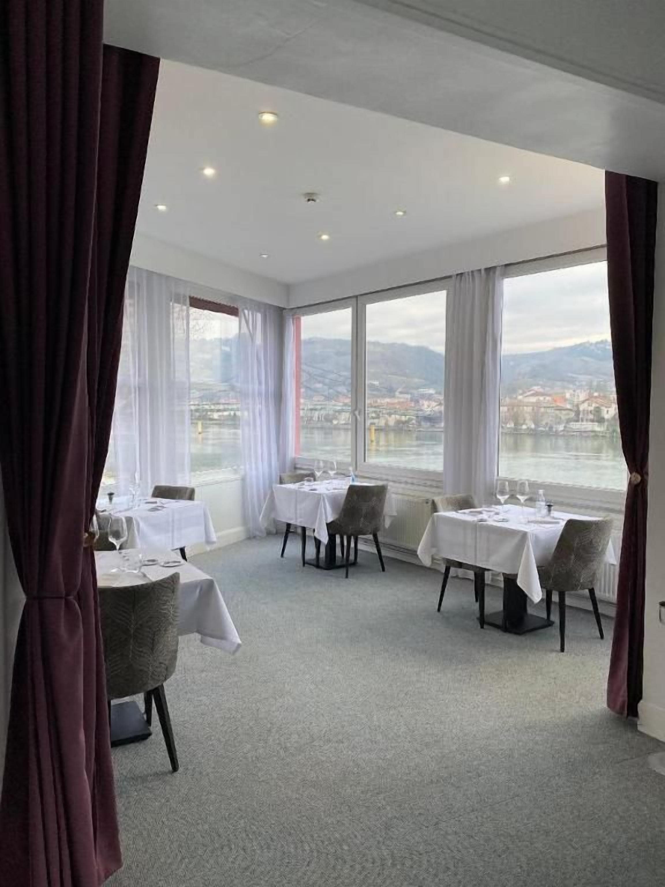 A dining area with tables set for two, large windows with a view of a body of water and mountains, and a carpeted floor.