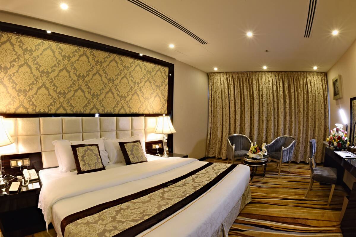 A hotel room with a large bed, two bedside tables with lamps, a desk with a chair, a large window with curtains, and a carpeted floor.