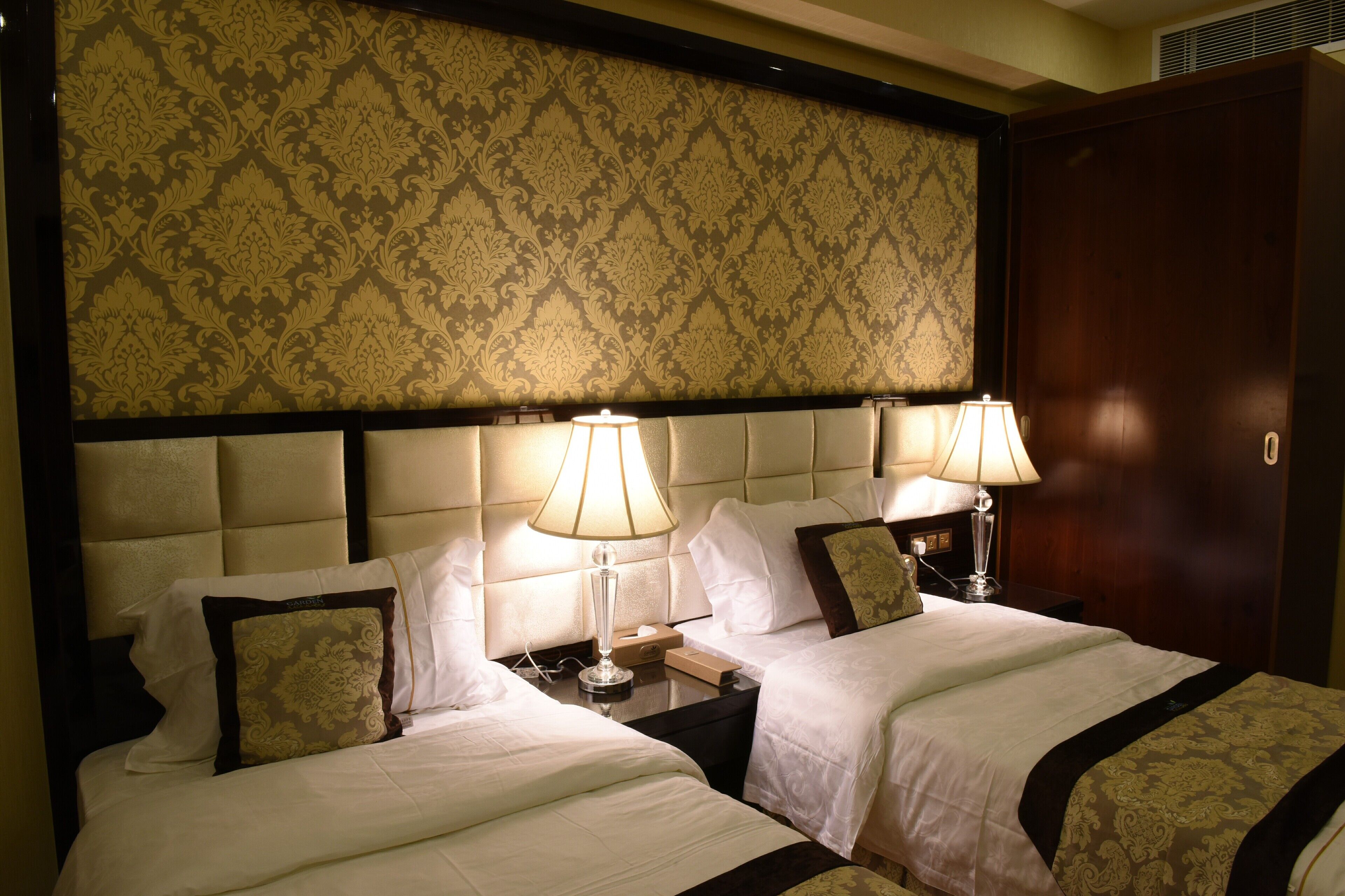 Habitación de hotel con dos camas, cada una con ropa blanca y almohadas decorativas, un cabecero grande con papel tapiz estampado y lámparas de noche.
