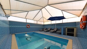 Una piscina interior con techo geométrico, paredes de azulejos azules, sillones de descanso y un flotador.