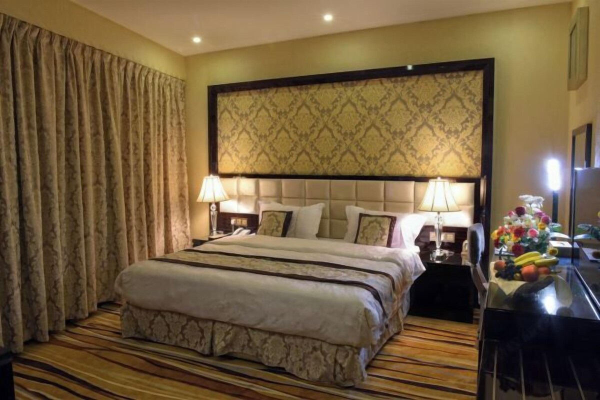 A hotel room with a large bed, two bedside lamps, a headboard with a patterned design, a wooden floor, and a nightstand with a fruit bowl.