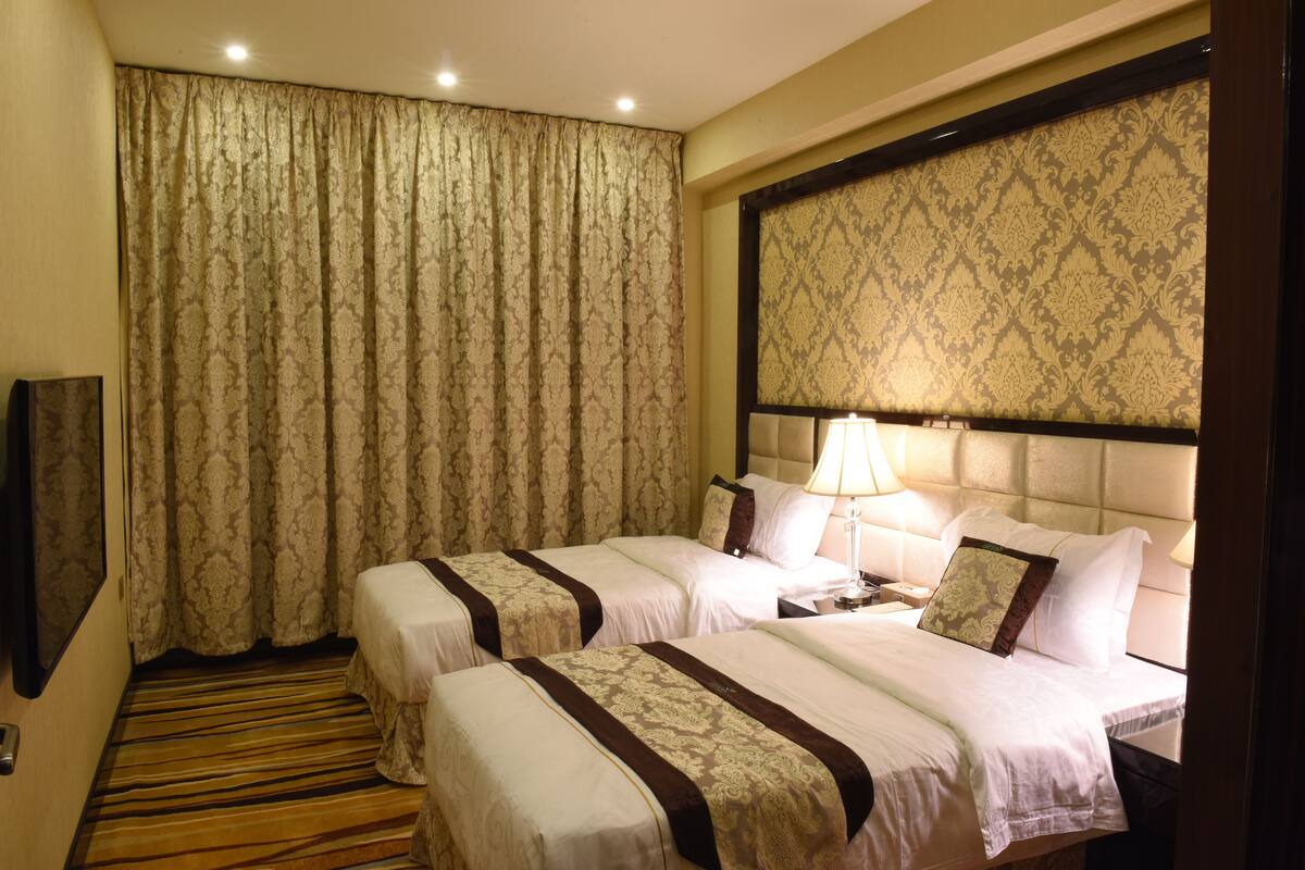 A hotel room with two beds, a large window with curtains, a headboard with a patterned design, and a bedside table with a lamp.