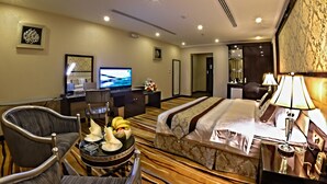 Habitación de hotel con una cama grande, un escritorio con televisor, una silla, una pequeña mesa con frutas y una lámpara.