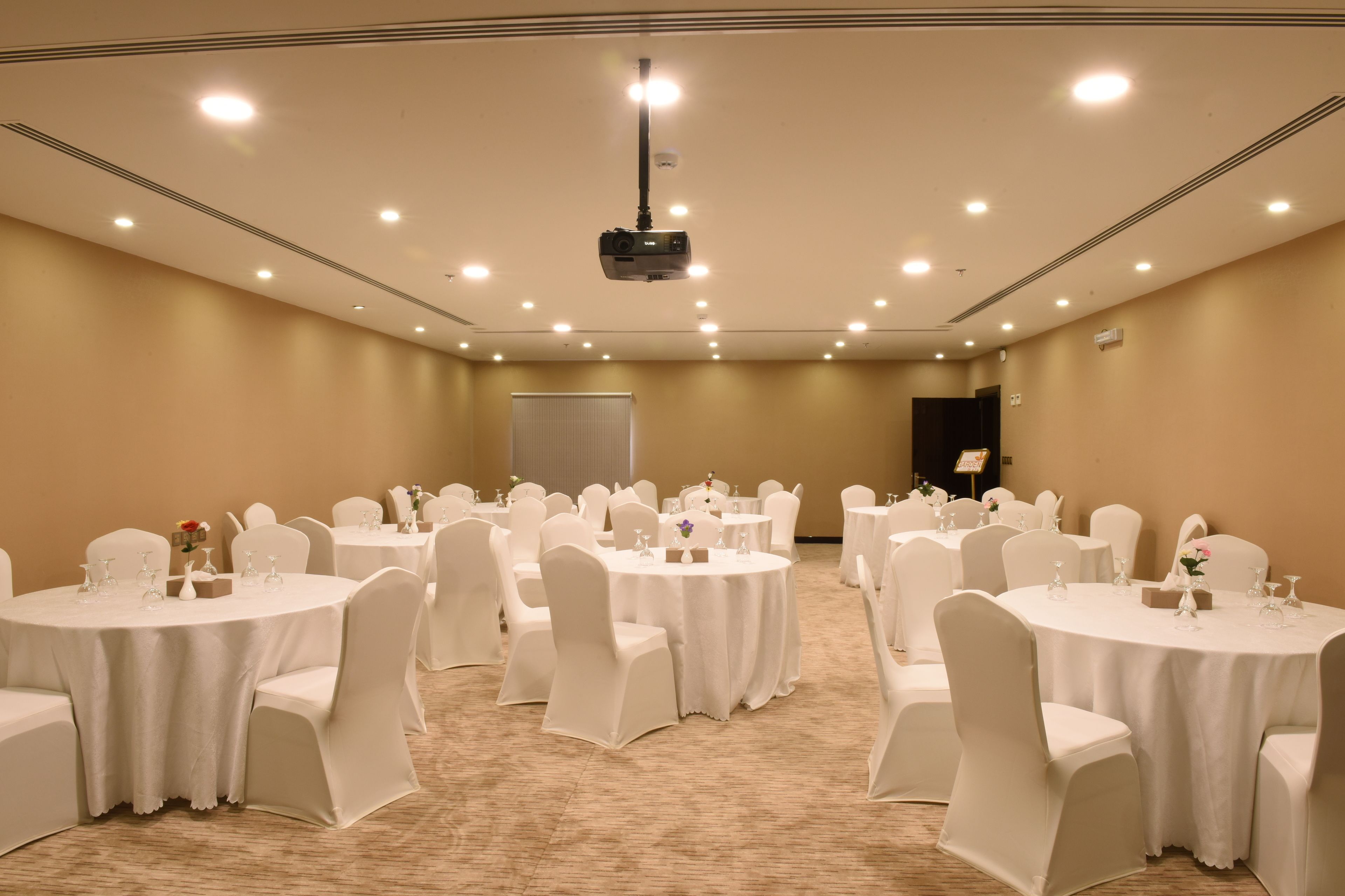 A conference room with multiple round tables, white chairs, and a projector mounted on the ceiling.