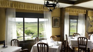 Un comedor con mesas dispuestas para una comida, ventanas amplias con cortinas y vistas a un paisaje verde.