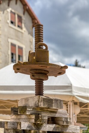 Un objeto metálico oxidado con una tuerca y una base de madera, frente a un edificio con techo rojo y una carpa blanca al fondo.