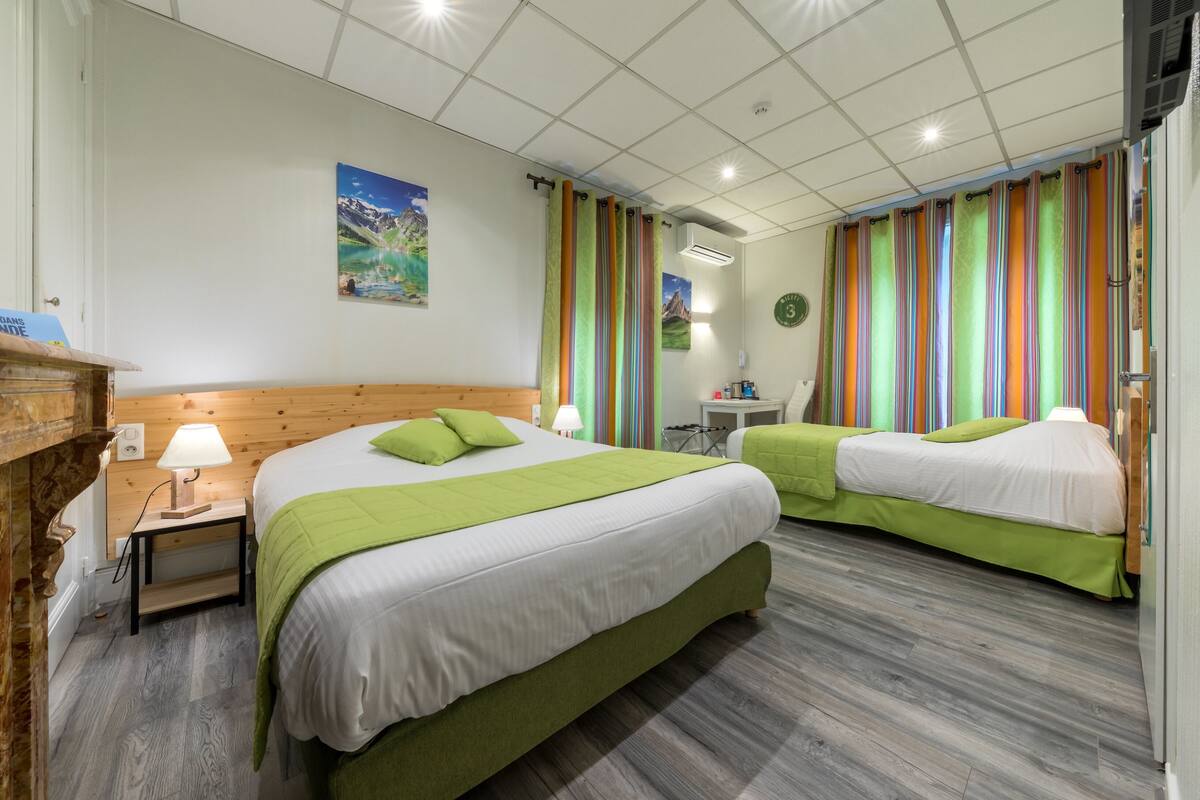 Habitación de hotel con dos camas, cabecera de madera, mesita de noche con lámpara, pequeña mesa con lámpara y cortinas de colores.